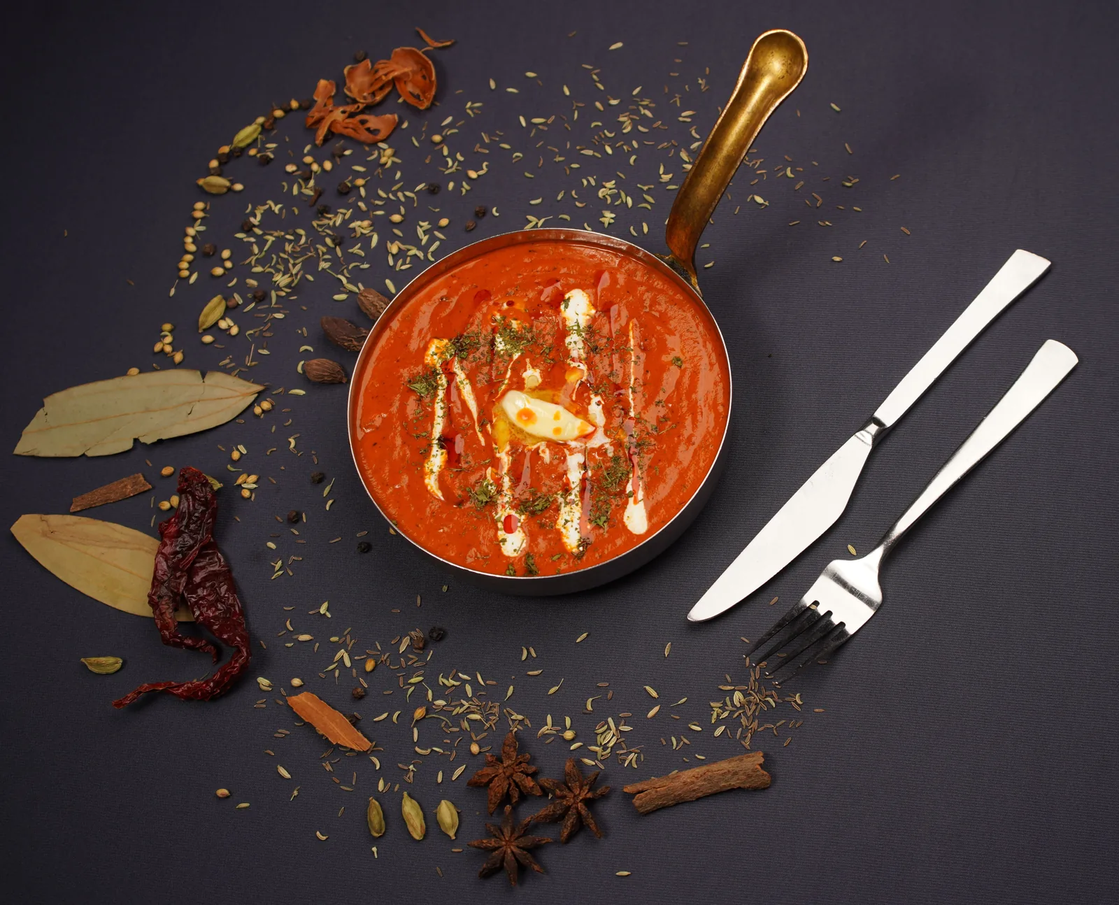 Creamy masala curry in a brass-handled pan surrounded by whole spices